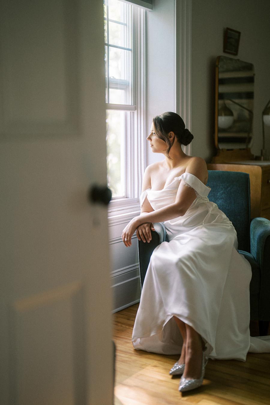 Elegant bride in an off-the-shoulder wedding gown sits on a blue armchair, gazing thoughtfully out a sunlit window, with a vintage mirror in the background.