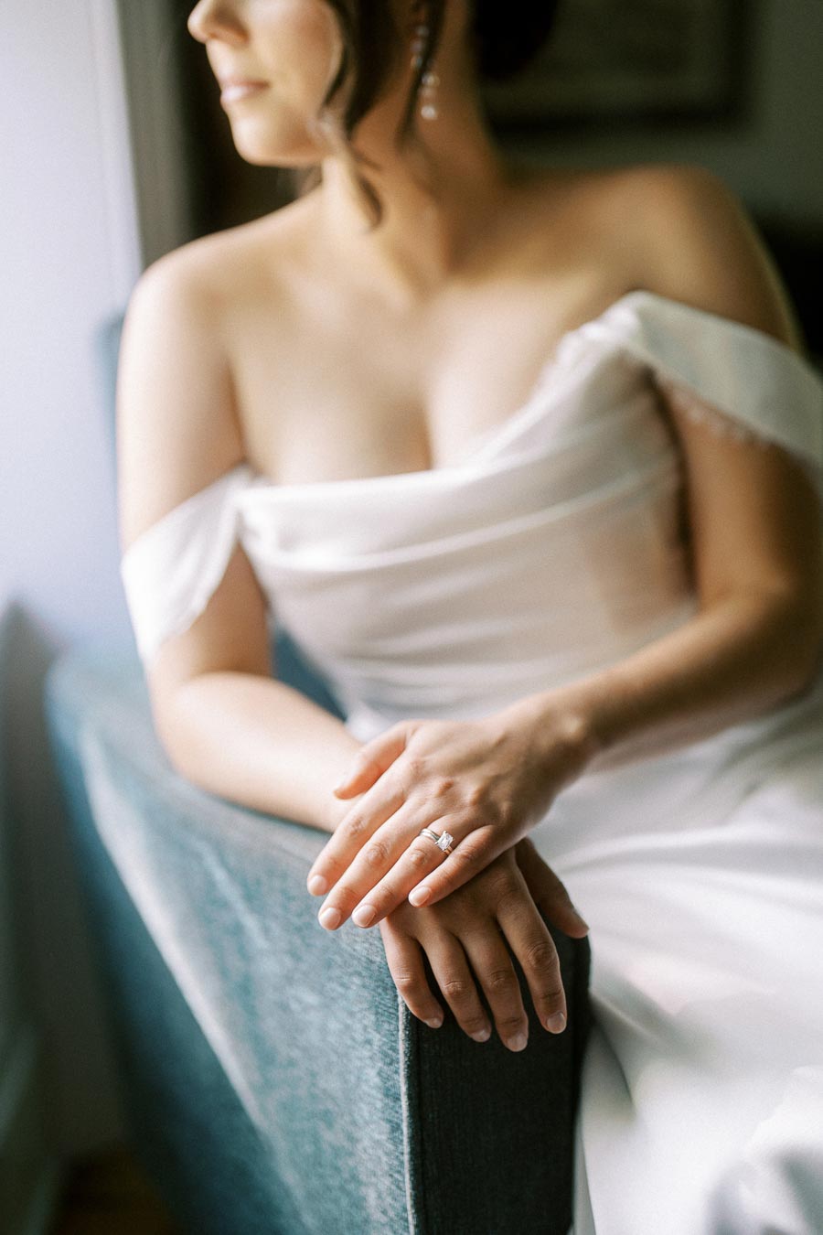 Elegant bride in an off-shoulder gown sitting on a chair, showcasing a beautiful diamond engagement ring against soft natural light.