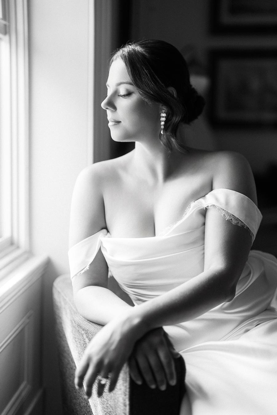 Black and white photo of a woman in an elegant off-shoulder dress sitting by a window, looking away with a serene expression and wearing pearl earrings.