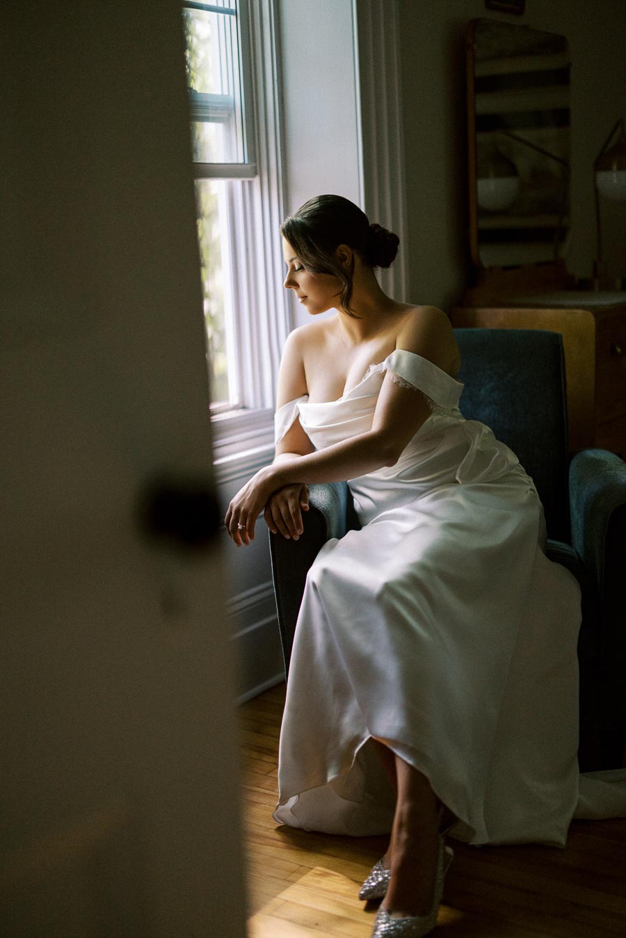 Elegant bride in white gown sitting by window, reflecting softly in natural light.