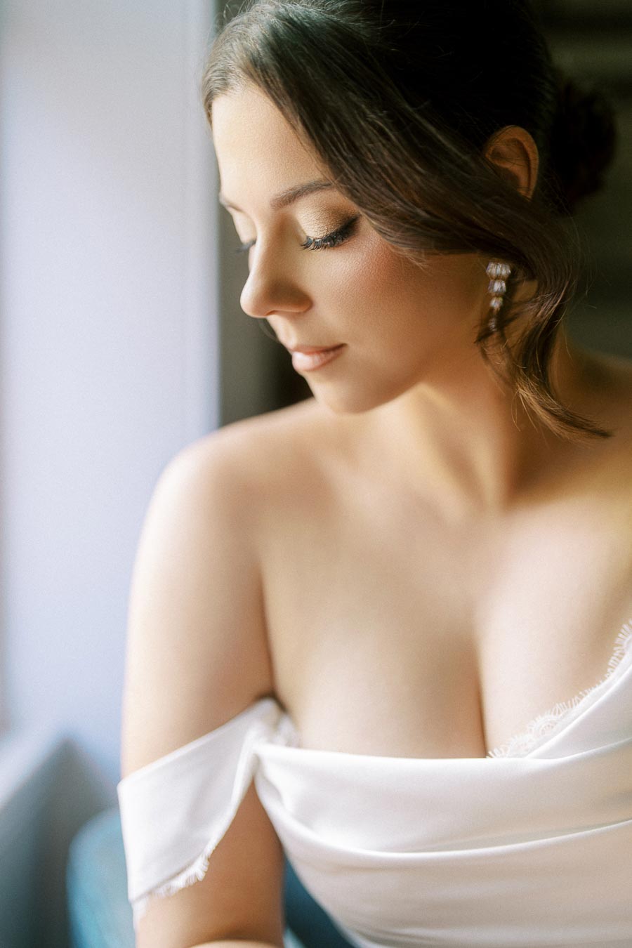 Elegant bride in a white off-shoulder dress gazing down, highlighting her soft makeup and pearl earrings.