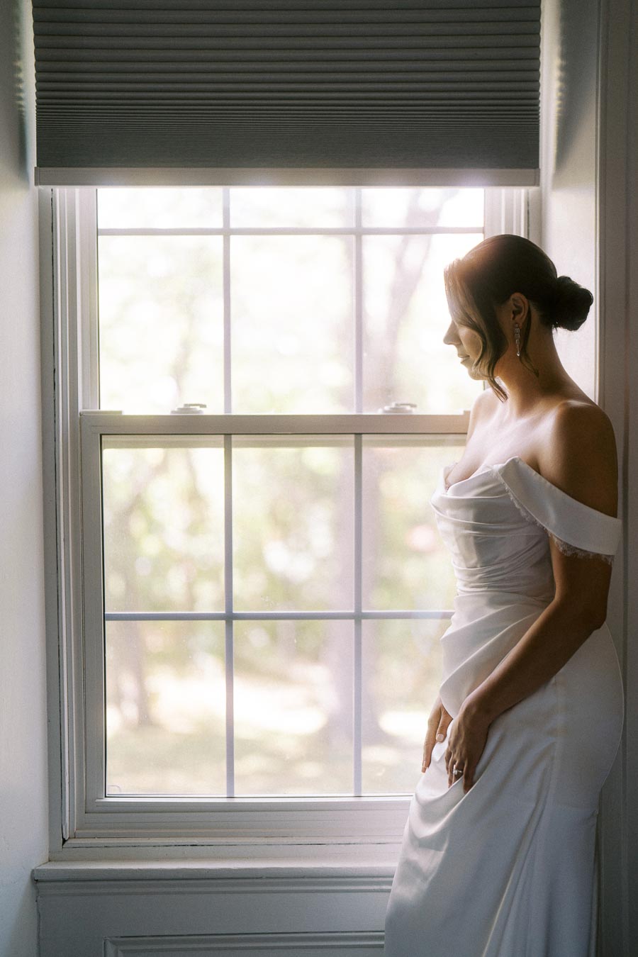 Bride in elegant off-shoulder wedding gown gazing out a bright window, creating a serene and romantic atmosphere.