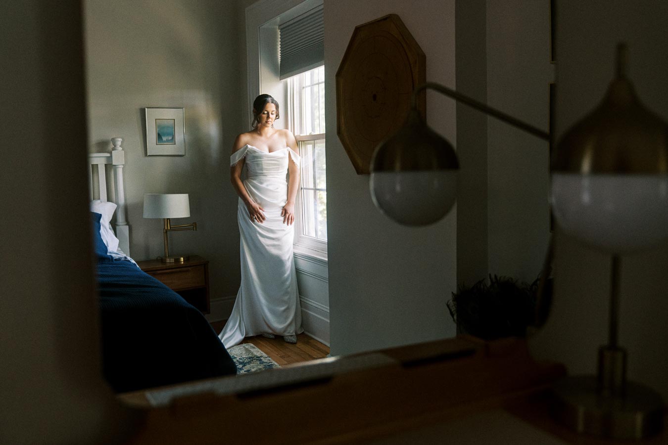 A bride in an elegant, off-shoulder white wedding dress stands by a window in a softly lit bedroom, featuring modern decor and a bedside lamp.