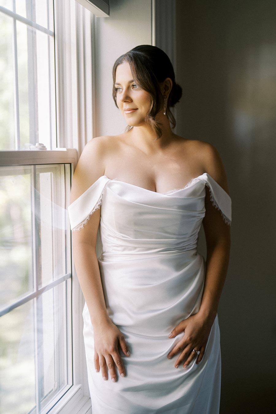A bride in an elegant off-the-shoulder white wedding dress poses by a window with soft, natural lighting.