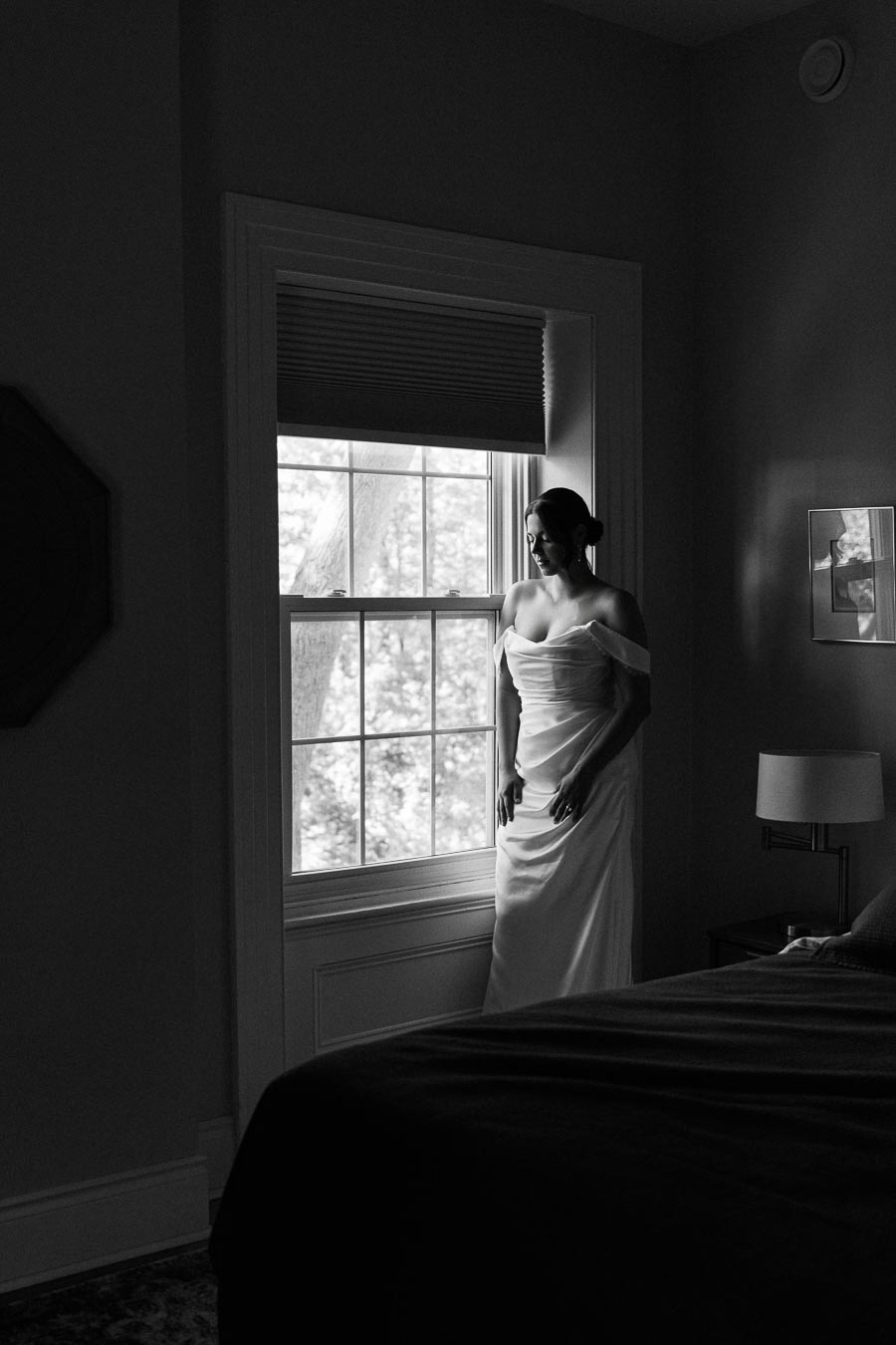 Black and white photo of a woman in an elegant off-shoulder dress standing by a large window, softly illuminated by natural light, creating a serene and reflective atmosphere in a dimly lit room.