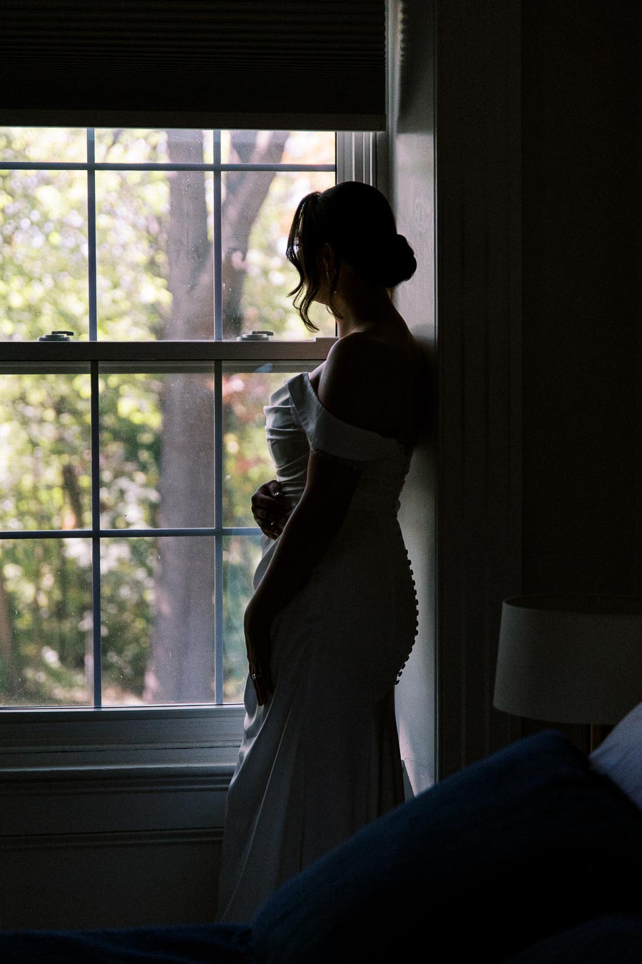 Bride in an elegant wedding dress standing in a softly lit room, gazing out of a window with a serene garden view.