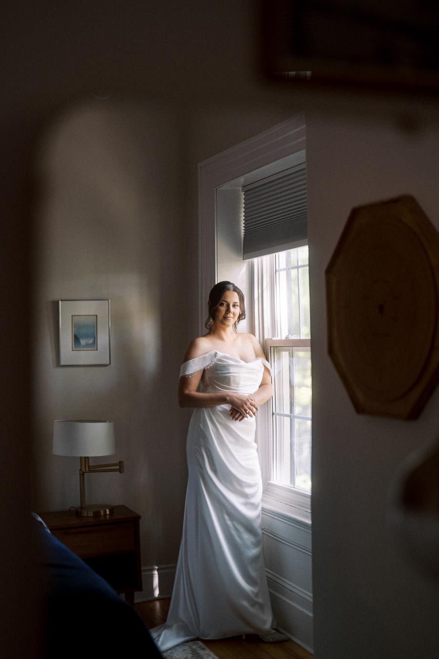 Bride in white wedding dress standing by a window, illuminated by soft natural light in a cozy room.