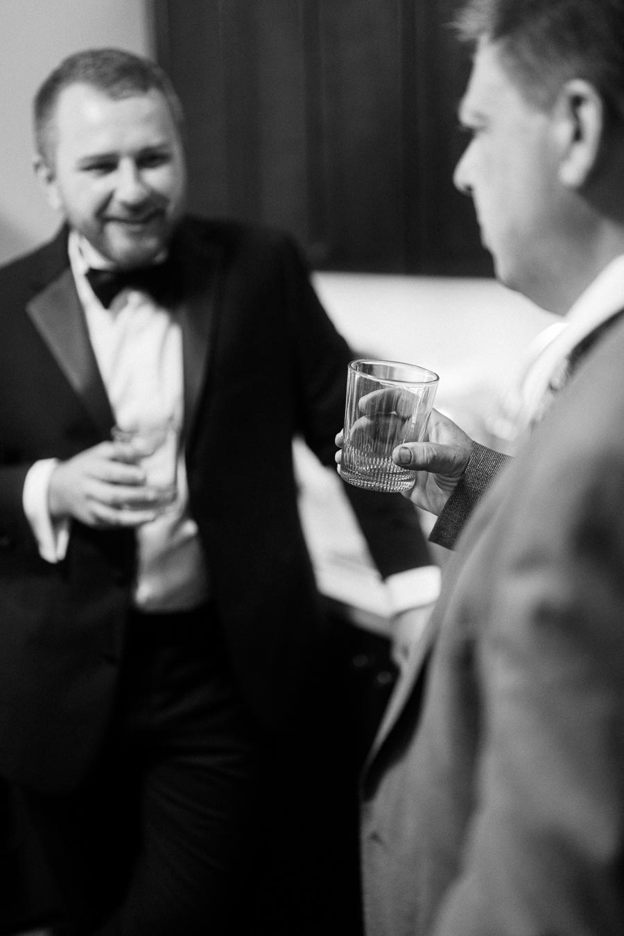 Black and white image of two men in formal suits holding whiskey glasses, engaged in a conversation at a social gathering.