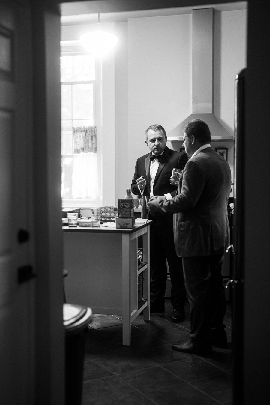 Two men in formal attire having a conversation in a kitchen, one holding a glass. The black-and-white photo features a small kitchen island with boxes and containers, under soft lighting from a nearby window.