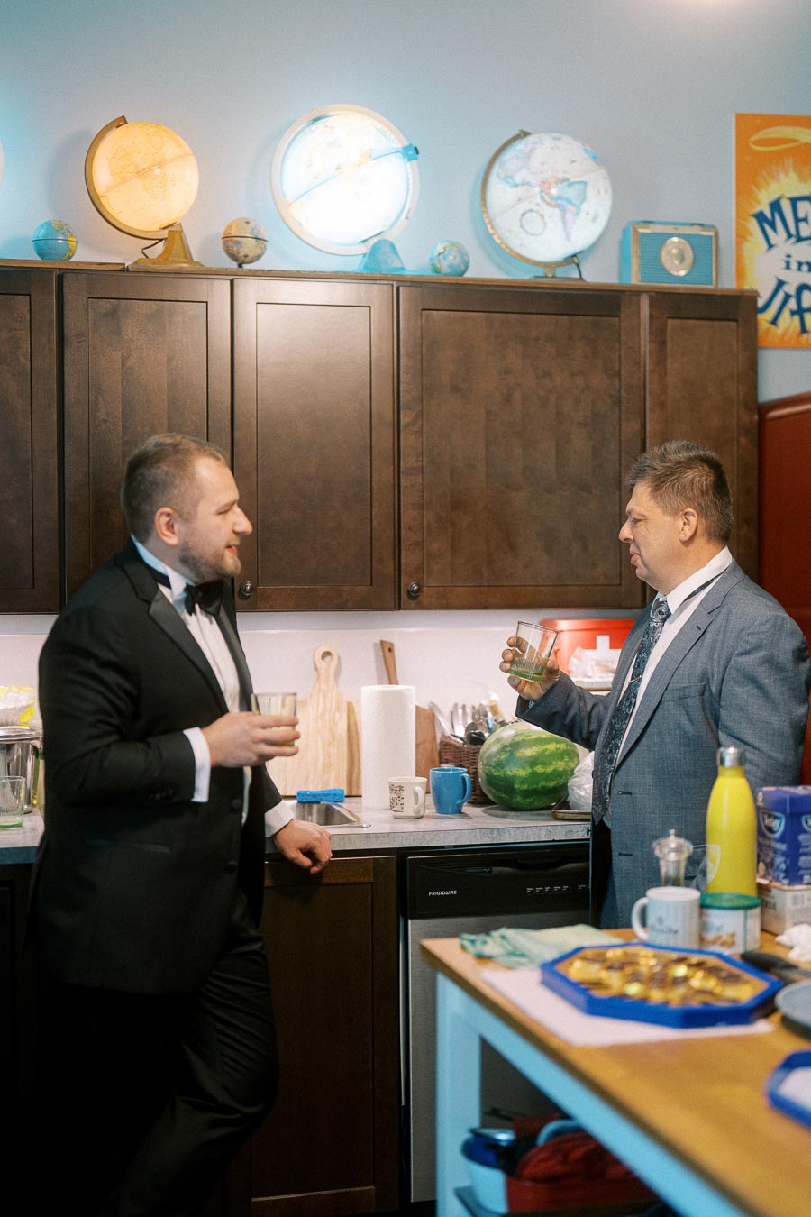 Two men in formal attire having a conversation in a kitchen decorated with globes, holding drinks near a counter with a watermelon and assorted items.