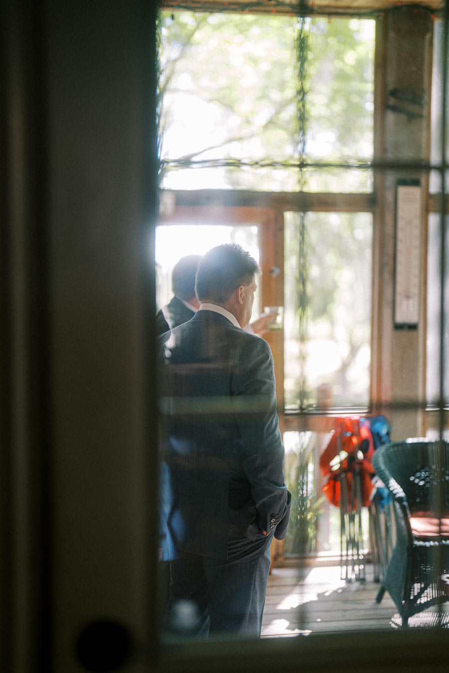 View through a glass window of two men in suits having a conversation in a sunlit room, with greenery visible outside.