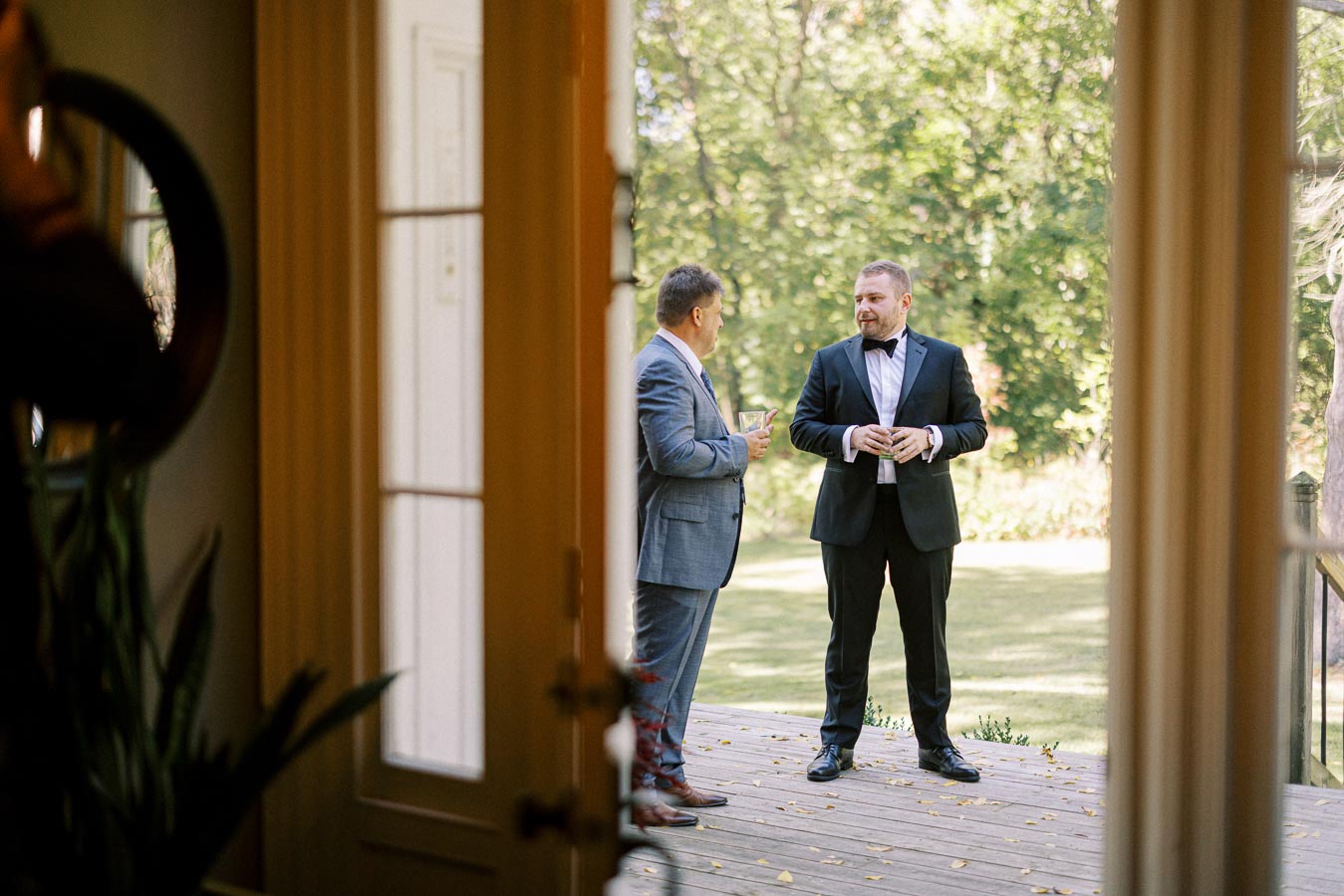 Two men in formal attire having a conversation on a wooden deck with a scenic green background, viewed through an open door.