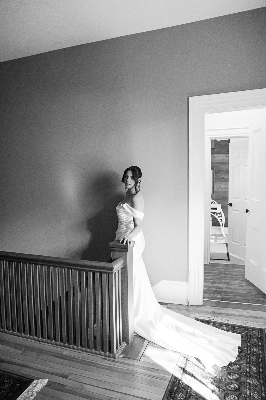 Black and white image of a woman in an elegant off-shoulder wedding dress posing gracefully on a staircase, with soft lighting creating a serene atmosphere.