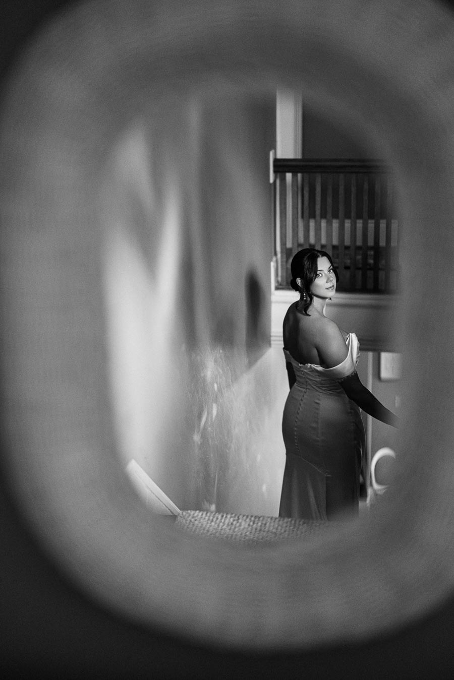 Black and white photo of a woman in an elegant dress, viewed through an oval frame, standing on a staircase and looking over her shoulder.