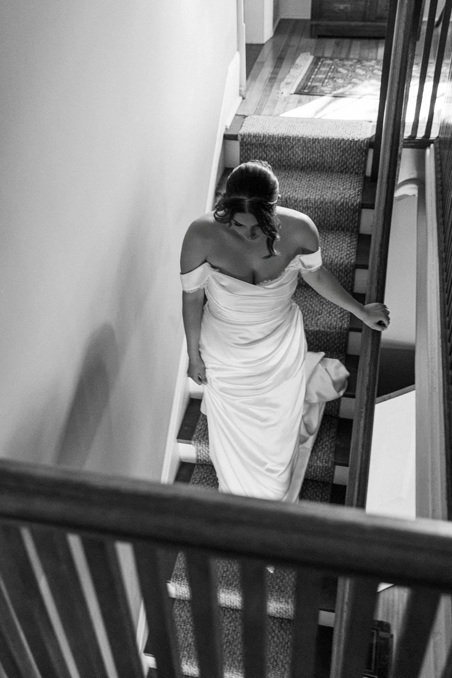 Bride in elegant off-shoulder wedding dress descending a staircase, captured in black and white for a vintage aesthetic.