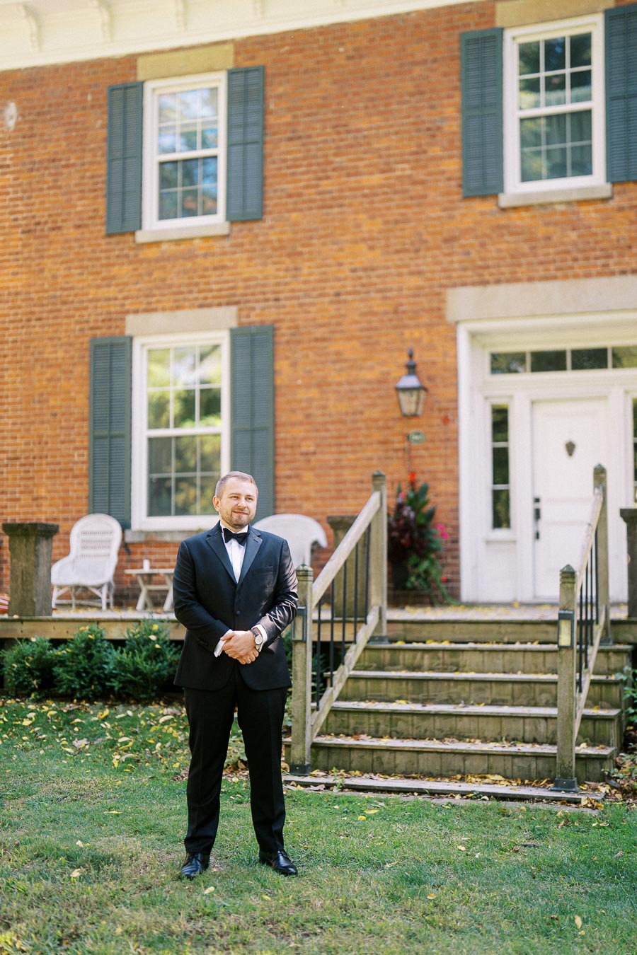 A man in a black tuxedo stands confidently in front of a brick house with blue shutters and a white door, surrounded by a lush green lawn and garden.
