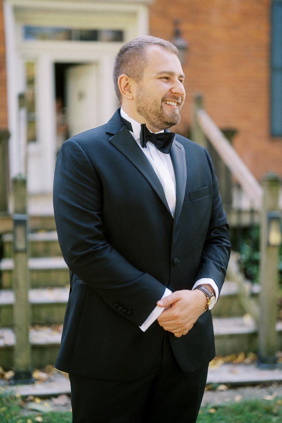 A man in a black tuxedo with a bow tie smiling outdoors in front of a brick building with stairs in the background.