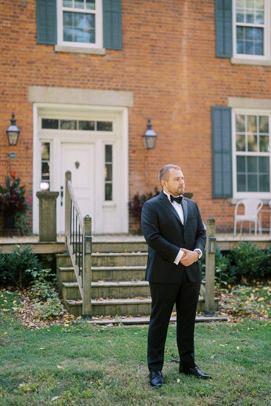 A man in a black tuxedo stands on a grassy lawn in front of a brick house with white trim and blue shutters, looking contemplative.