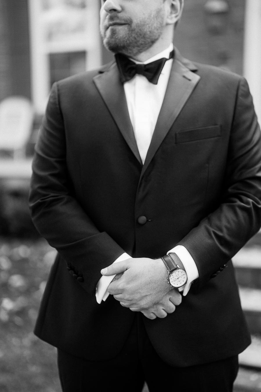 A man in a formal black tuxedo and bow tie, standing outdoors with hands clasped, wearing a stylish wristwatch.