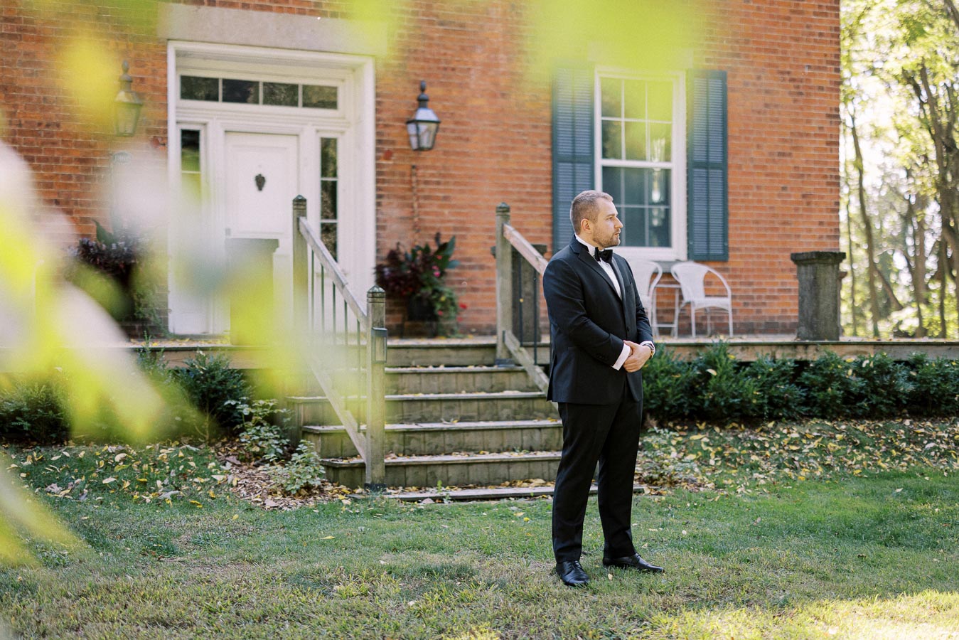 A man in a formal black tuxedo stands on a grassy lawn in front of a brick house with white trim and green shutters, surrounded by leafy greenery.