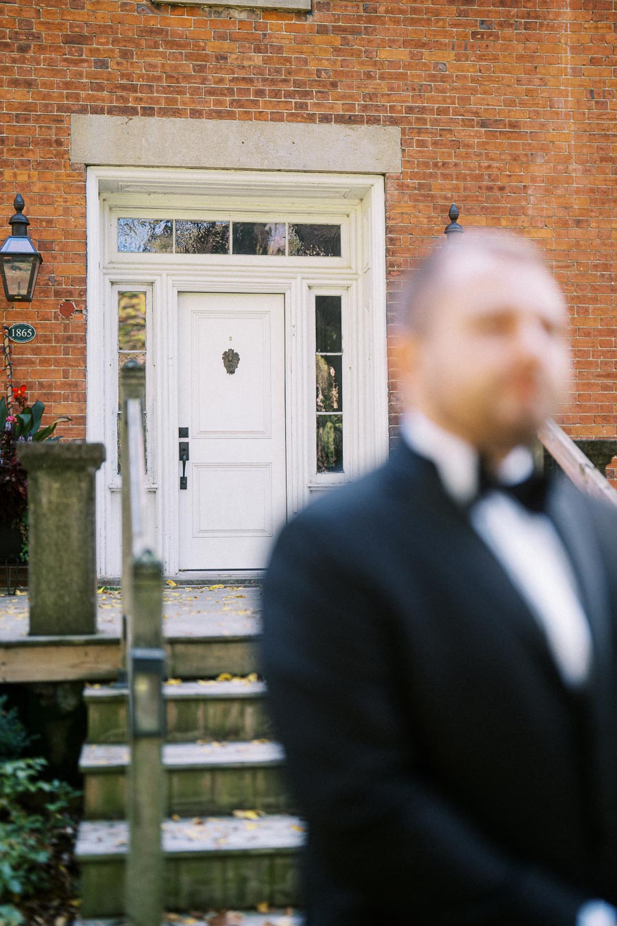 Blurred man in a black tuxedo standing in front of a historic brick building, focusing on an ornate white front door with vintage lanterns.