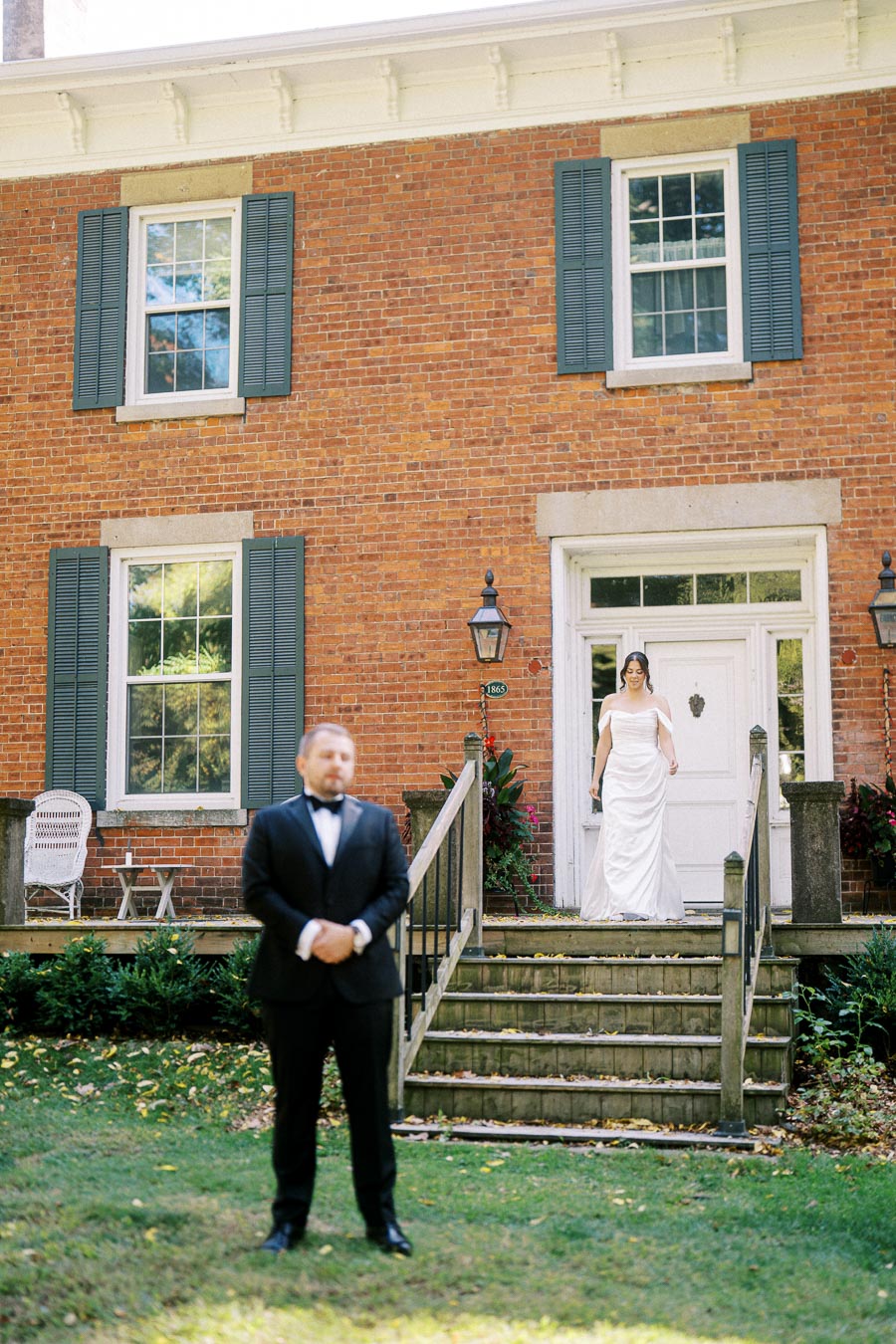 A bride in a white gown descends steps towards a groom in a tuxedo for a wedding first look outside a historic red brick building with blue shutters.