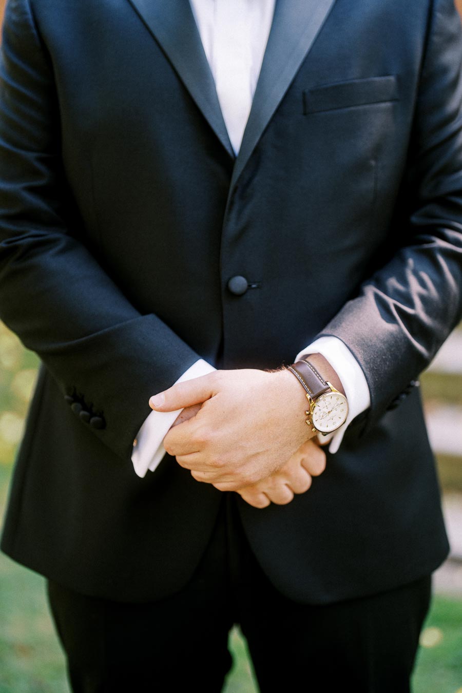 A man in a black suit and white shirt stands with hands together, showcasing a classic leather-strap wristwatch, outdoors on a sunny day.