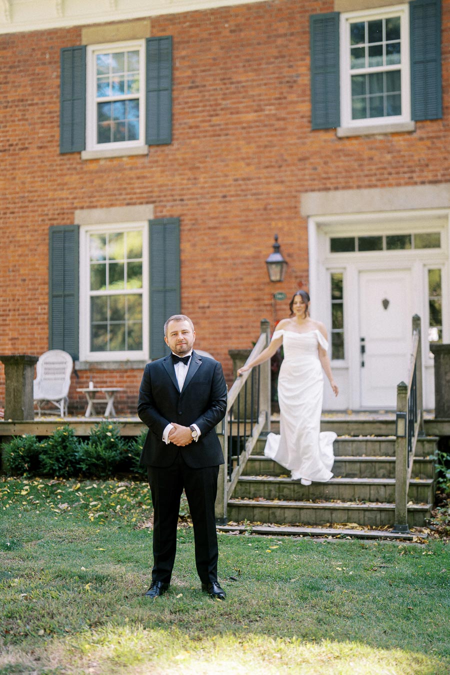 A bride and groom in formal attire pose in front of a classic brick house, with the bride walking down steps towards the groom standing in the garden.