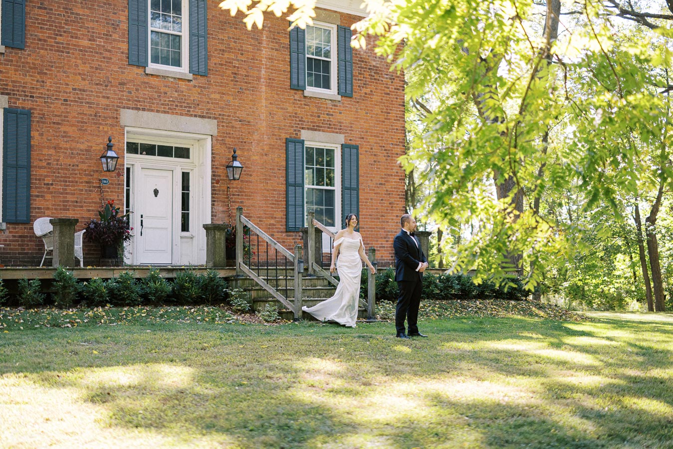 Bride approaching groom for first look in front of historic brick house with green shutters and lush garden setting.