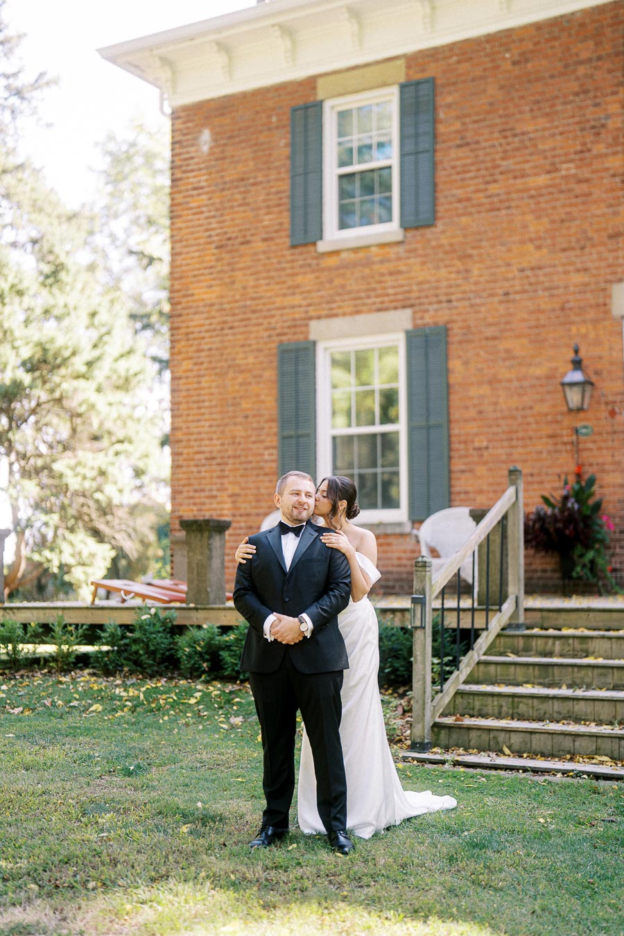 Bride and groom in elegant attire share a romantic moment in front of a historic brick building with greenery and steps.
