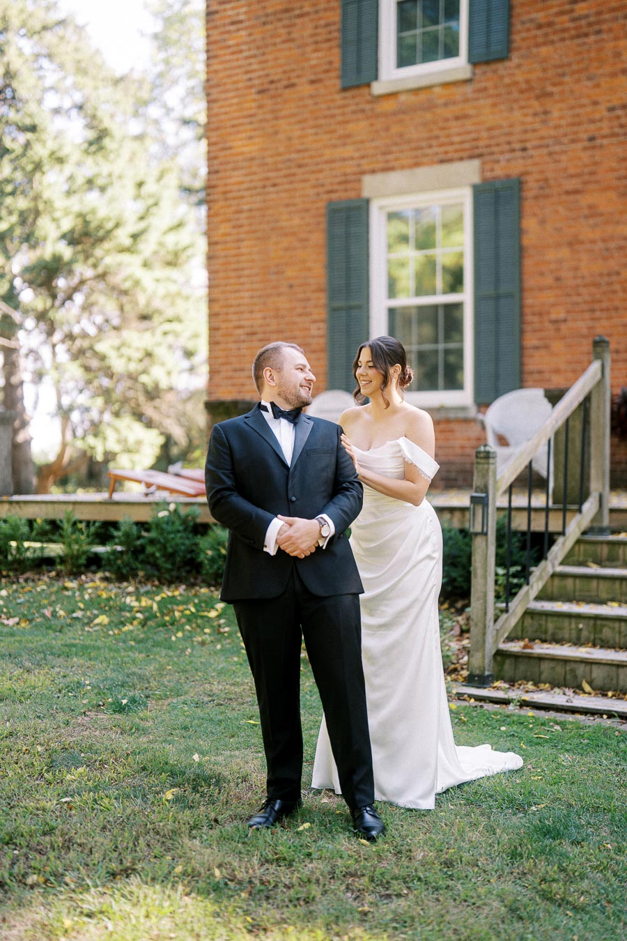 A bride and groom standing together in a garden next to a brick house on their wedding day; the groom is wearing a classic black tuxedo and the bride is in an elegant white wedding gown, both smiling and looking at each other.