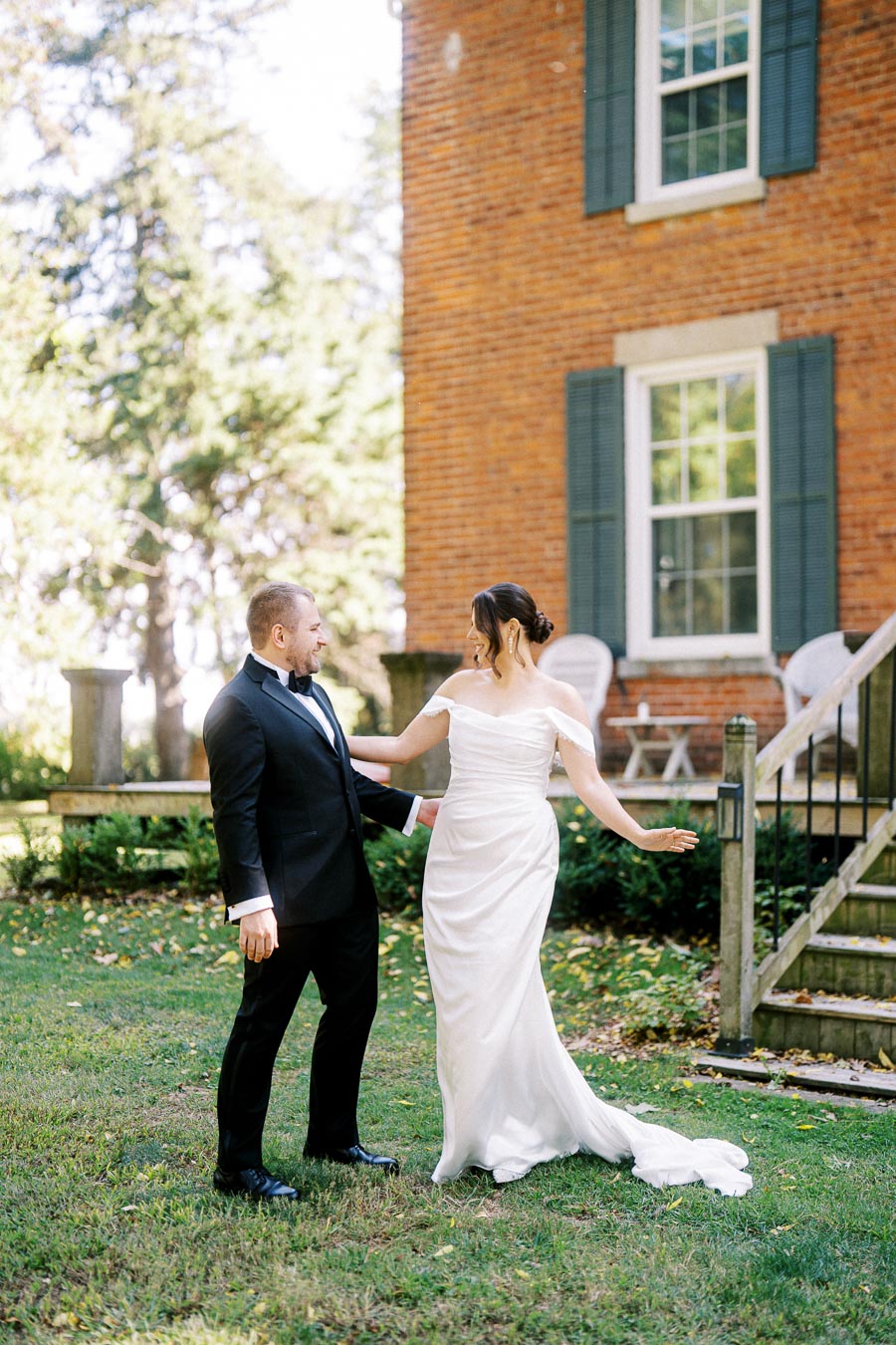A bride and groom smiling and embracing outdoors on their wedding day, standing in front of a rustic brick house with green window shutters, amidst a lush garden setting.