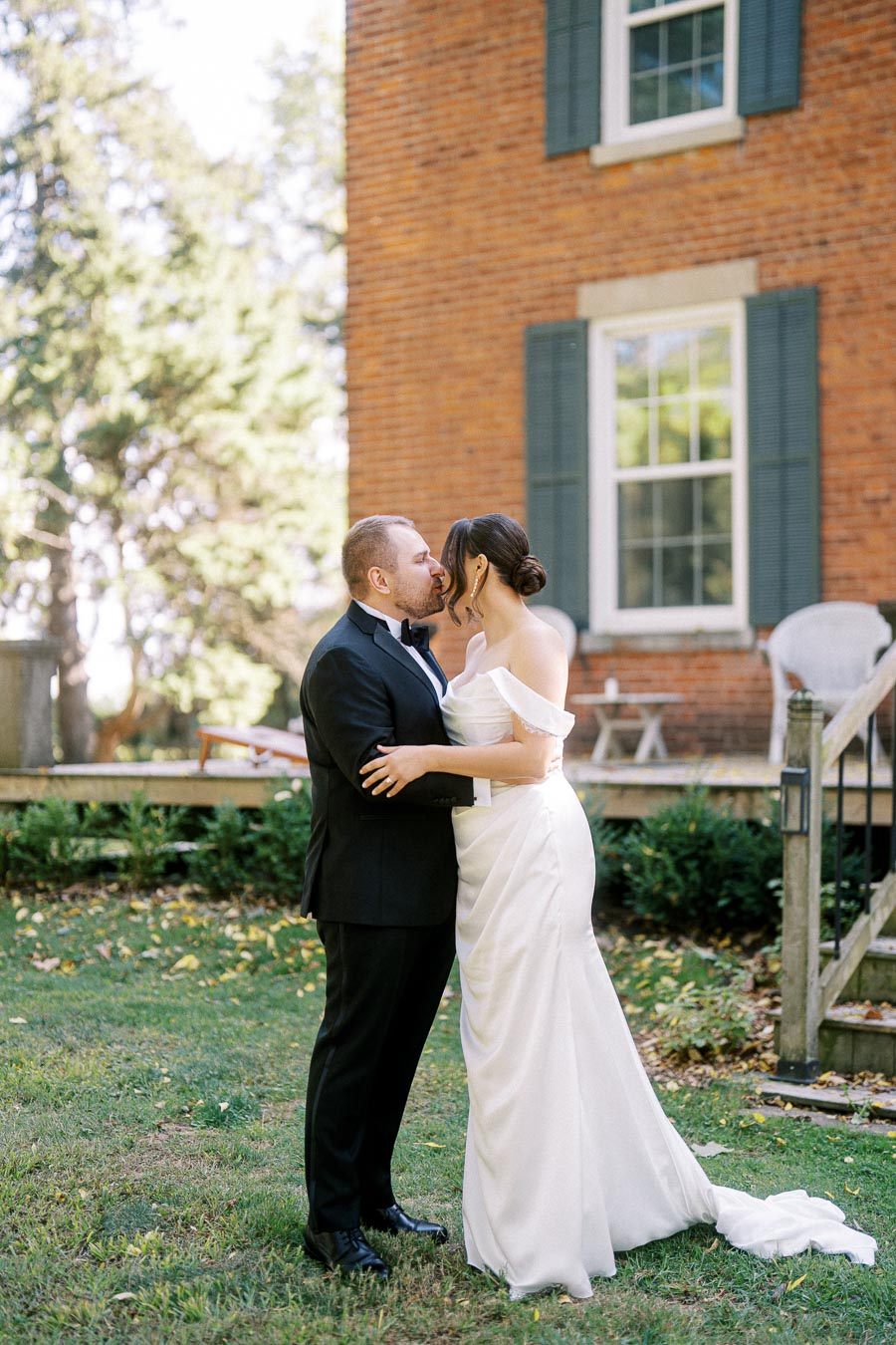 A bride and groom share a kiss in front of a rustic brick house, with greenery and trees in the background, creating a romantic outdoor wedding scene.