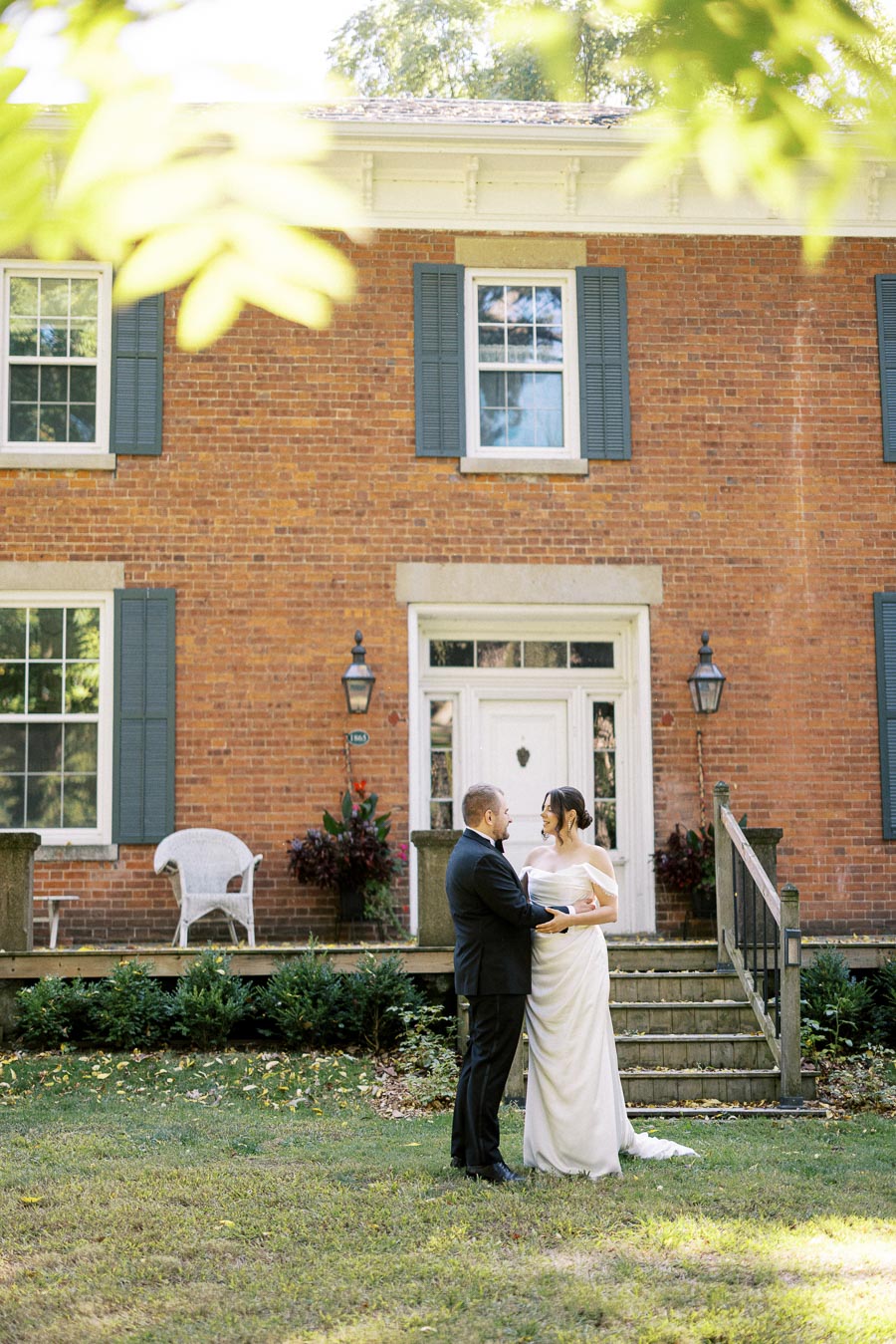 A bride and groom standing together in front of a charming brick house with blue shutters, surrounded by greenery on a sunny day.