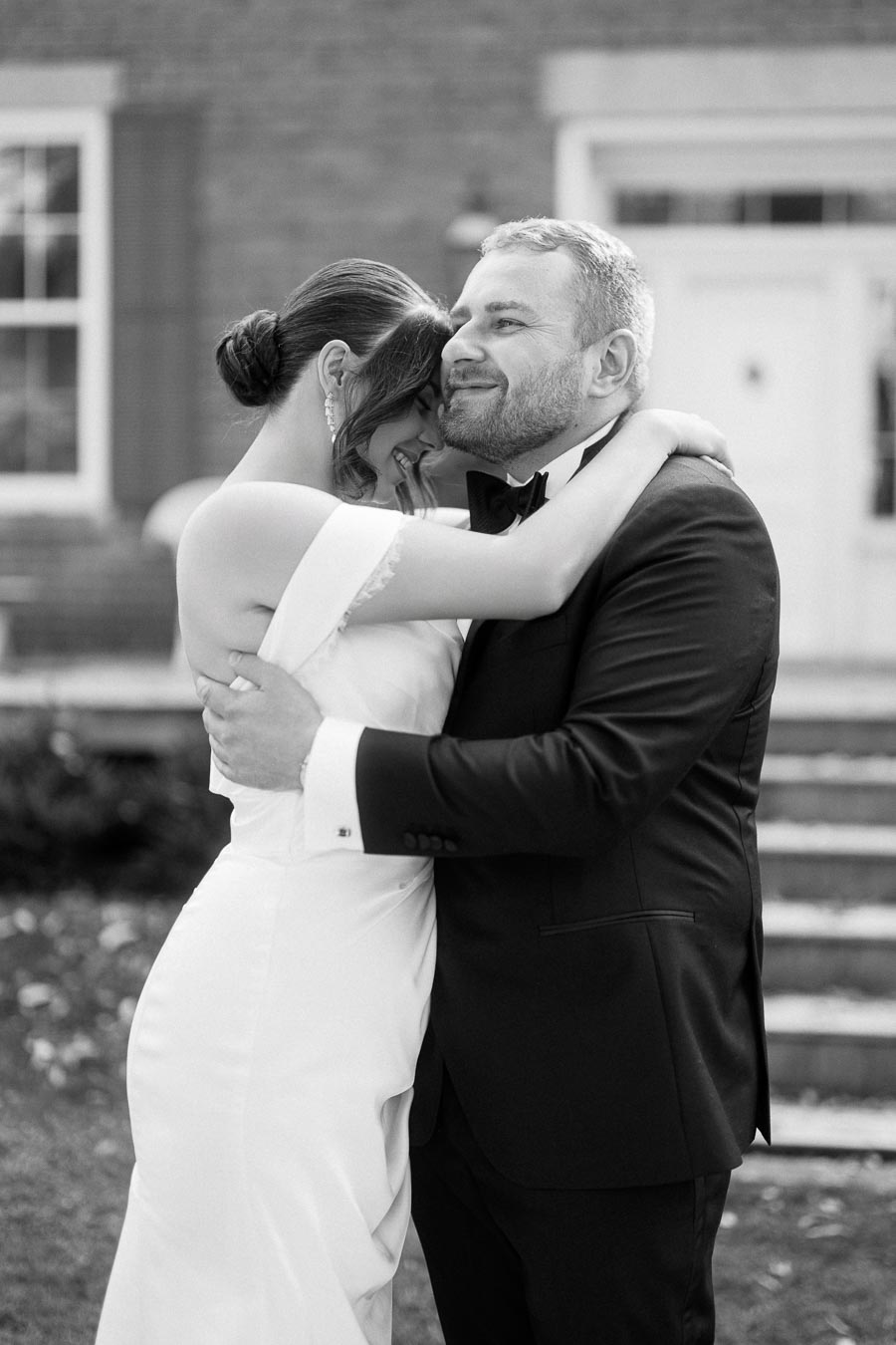 Black and white photo of a joyful couple embracing on their wedding day, with the bride in a white dress and the groom in a black suit, capturing a moment of love and happiness outside a building.