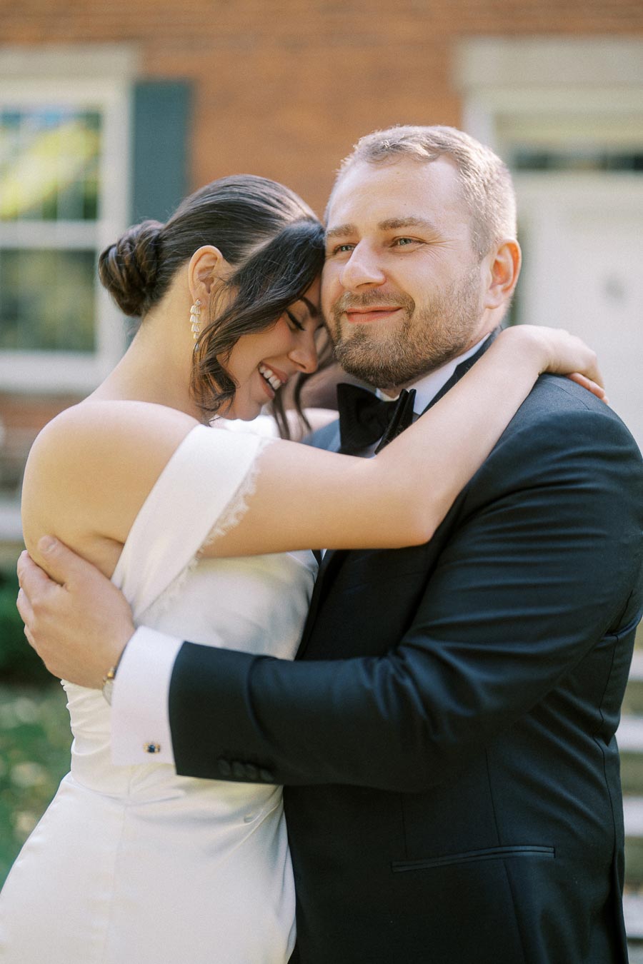 A joyful couple embracing on their wedding day, the bride in an elegant white dress and the groom in a black tuxedo, smiling in front of a brick building.