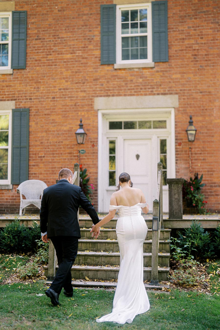 Bride and groom holding hands as they ascend stairs towards a charming brick house with white trim, surrounded by greenery and decorative lanterns, capturing a romantic wedding moment.