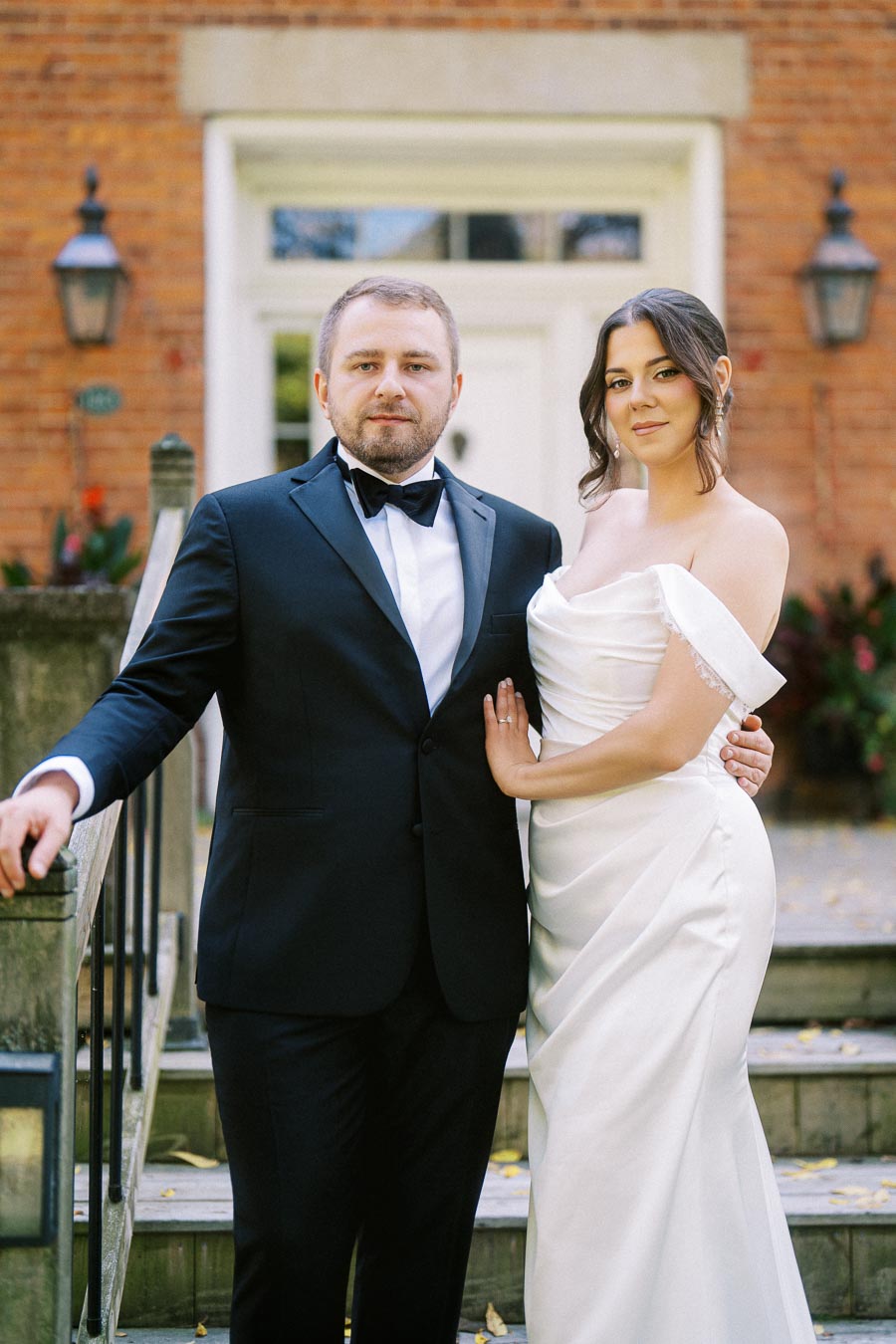 A couple posing for a formal wedding portrait in front of a brick building, with the man in a tuxedo and the woman in an elegant white wedding gown.