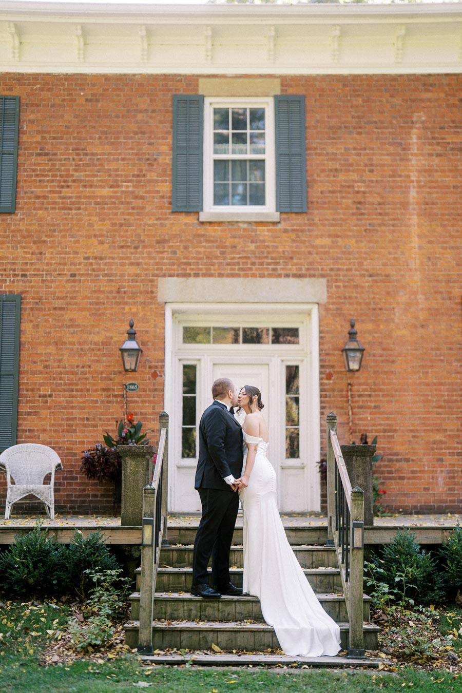 Elegant wedding couple sharing a romantic moment on a classic brick house staircase, bride in a flowing white gown and groom in a black tuxedo, surrounded by lush greenery and vintage decor.