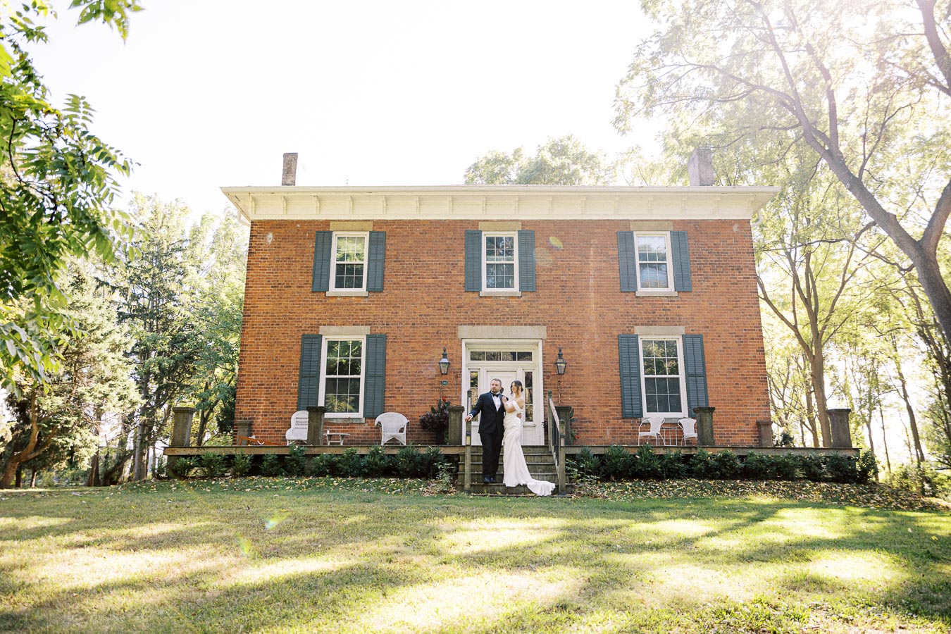 Wedding couple standing on the steps of a charming brick house surrounded by greenery and sunlight.