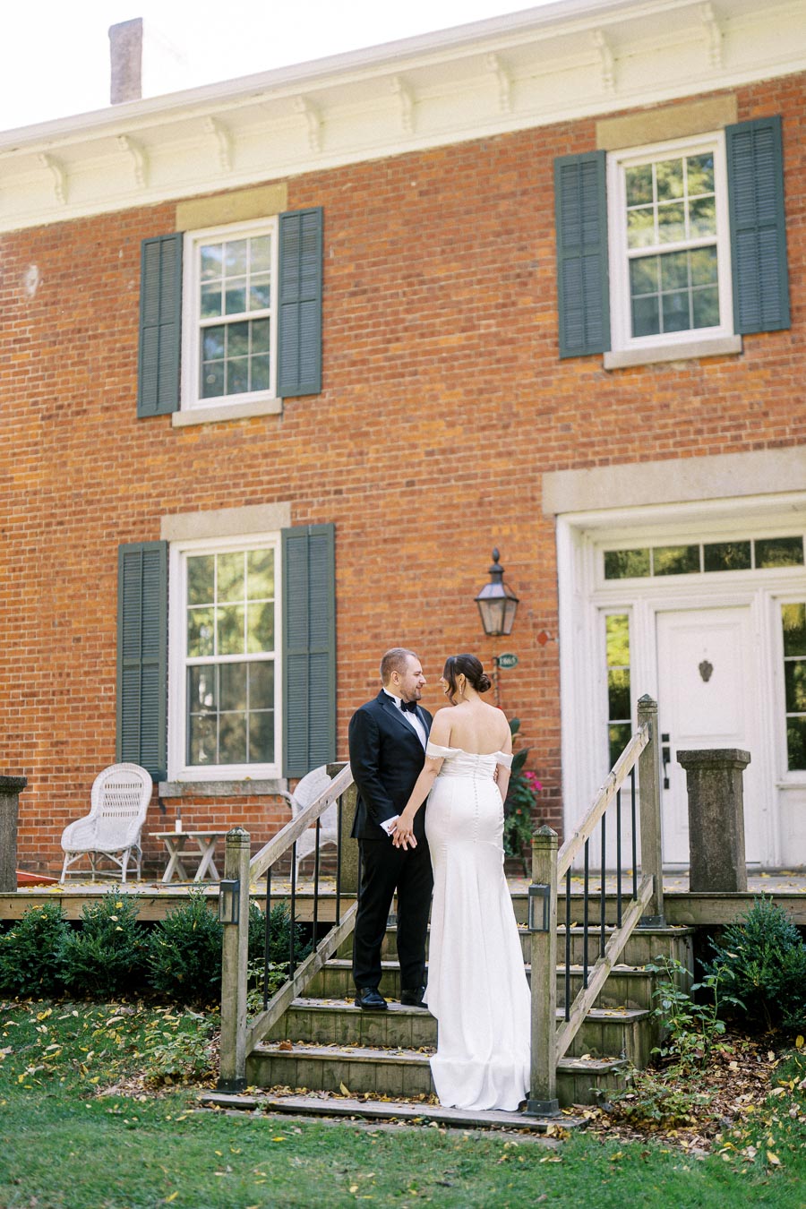 Newlywed couple holding hands on garden steps in front of rustic brick home with blue shutters and white trim, showcasing elegant wedding attire.