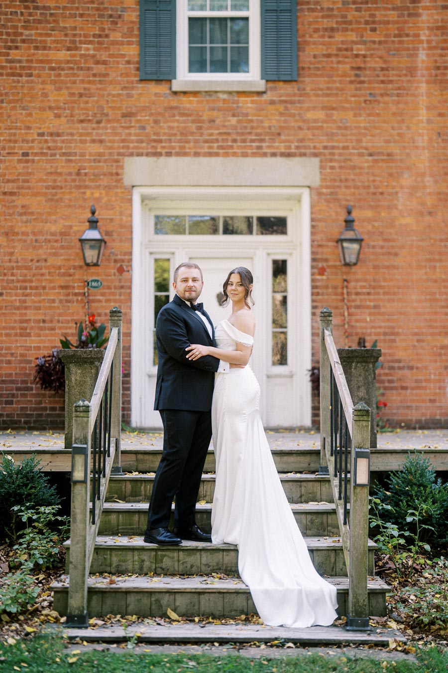Bride and groom in elegant wedding attire standing on rustic wooden steps against a brick house backdrop with green foliage.