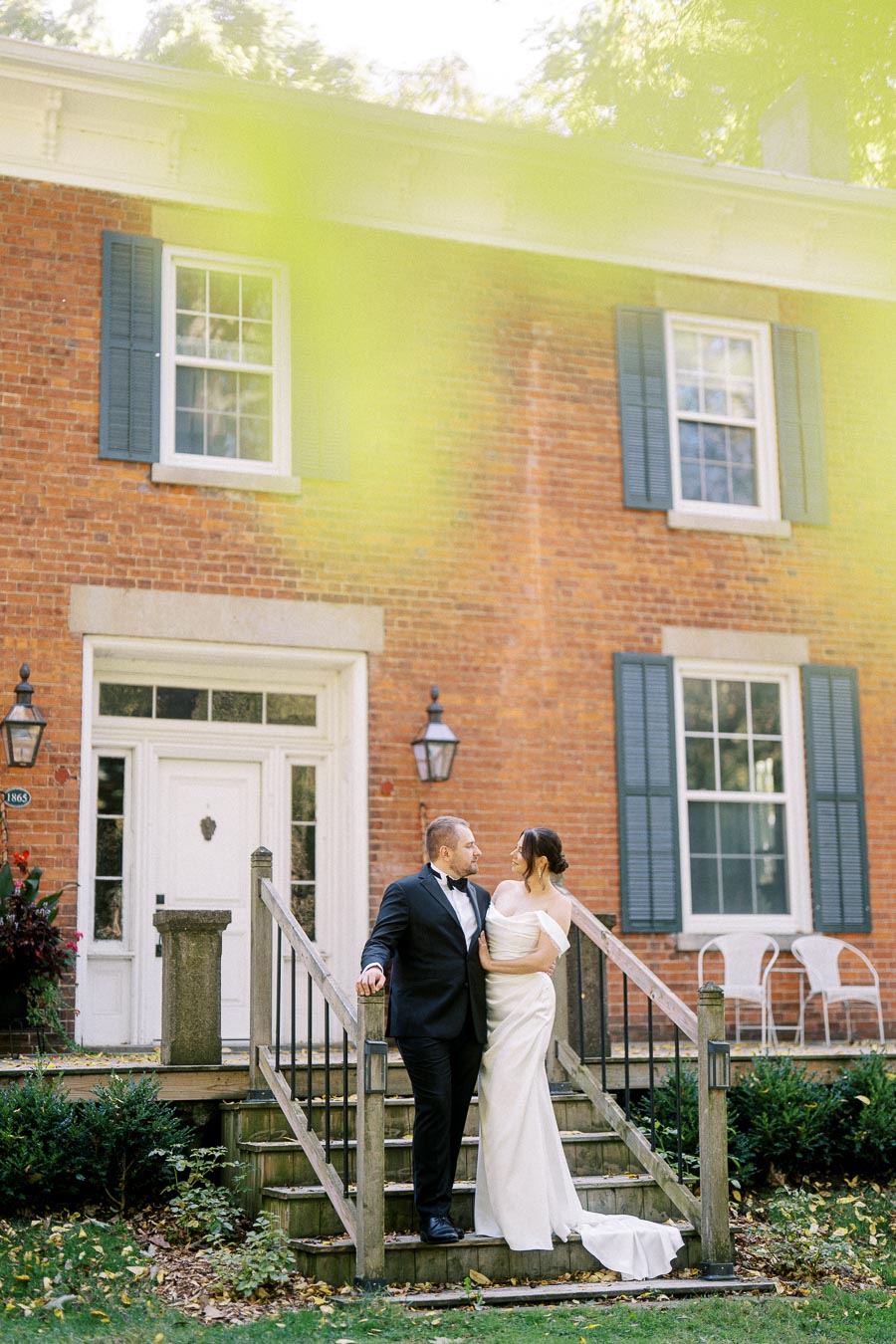 A newlywed couple poses lovingly on the steps of a historic brick house with blue shutters, surrounded by lush greenery.
