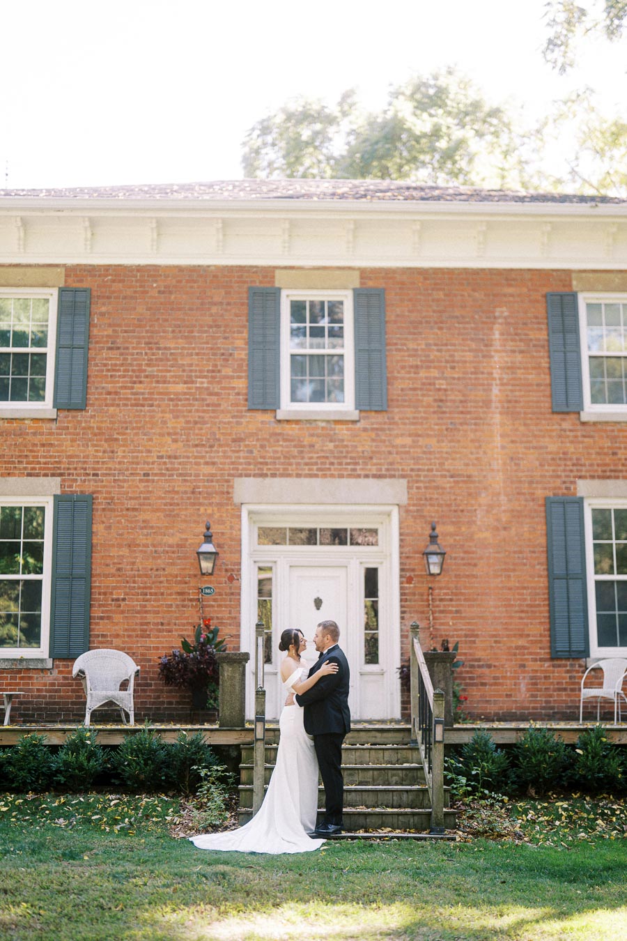 A newlywed couple embracing in front of a charming historic brick building with blue shutters and a classic wooden door, surrounded by lush greenery.