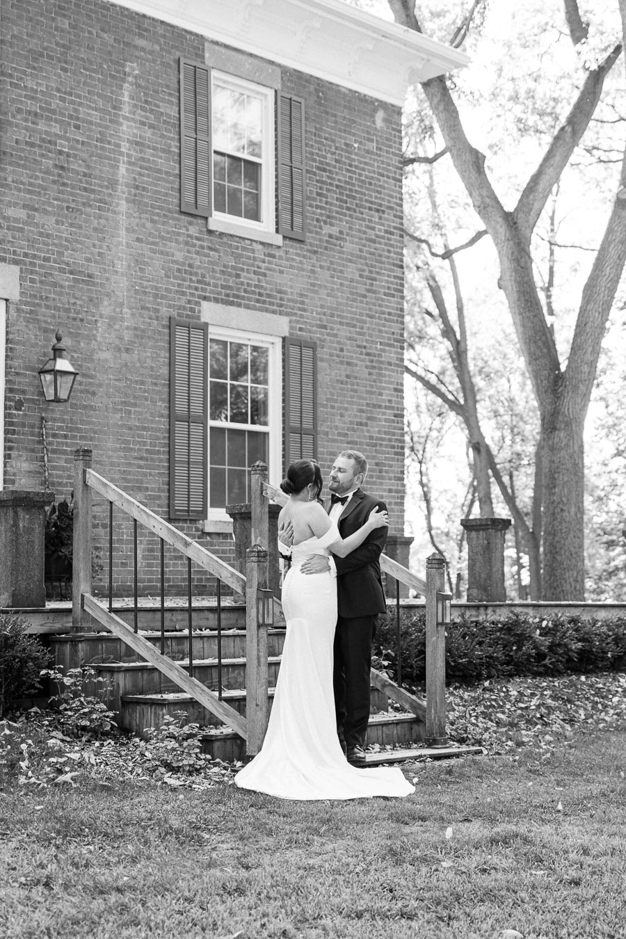 Black and white image of a couple embracing in front of a historic brick house. The woman is in a long, elegant white wedding dress and the man in a classic black suit. The scene captures a romantic moment on a wooden staircase surrounded by natural greenery and large trees, evoking a timeless, vintage feel.
