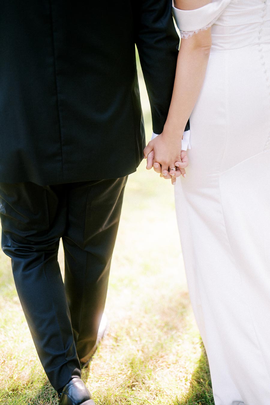 A bride and groom holding hands, walking on grass, with the bride wearing a white dress and the groom in a black suit, symbolizing love and commitment.