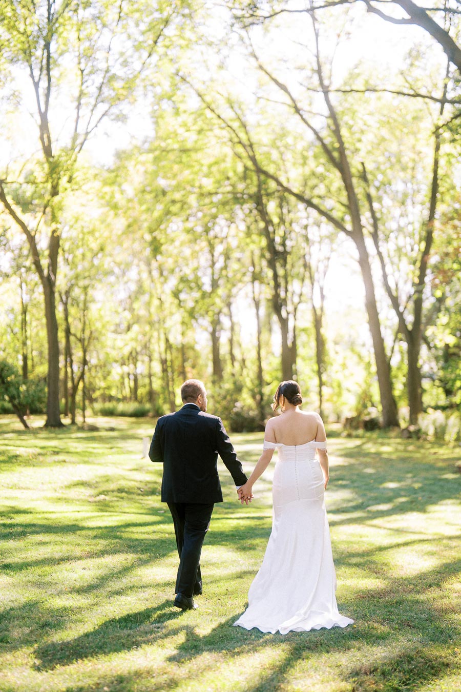Couple walking hand in hand through a sunlit forest, bride wearing a white wedding dress and groom in a black suit, surrounded by lush green trees.