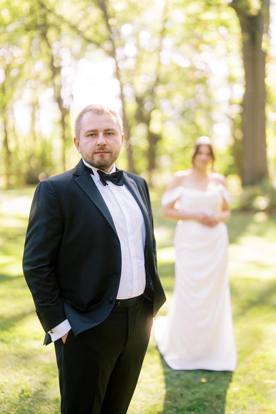 Groom in formal black tuxedo and bow tie standing in a sunlit garden with blurred bride in white wedding dress in the background.