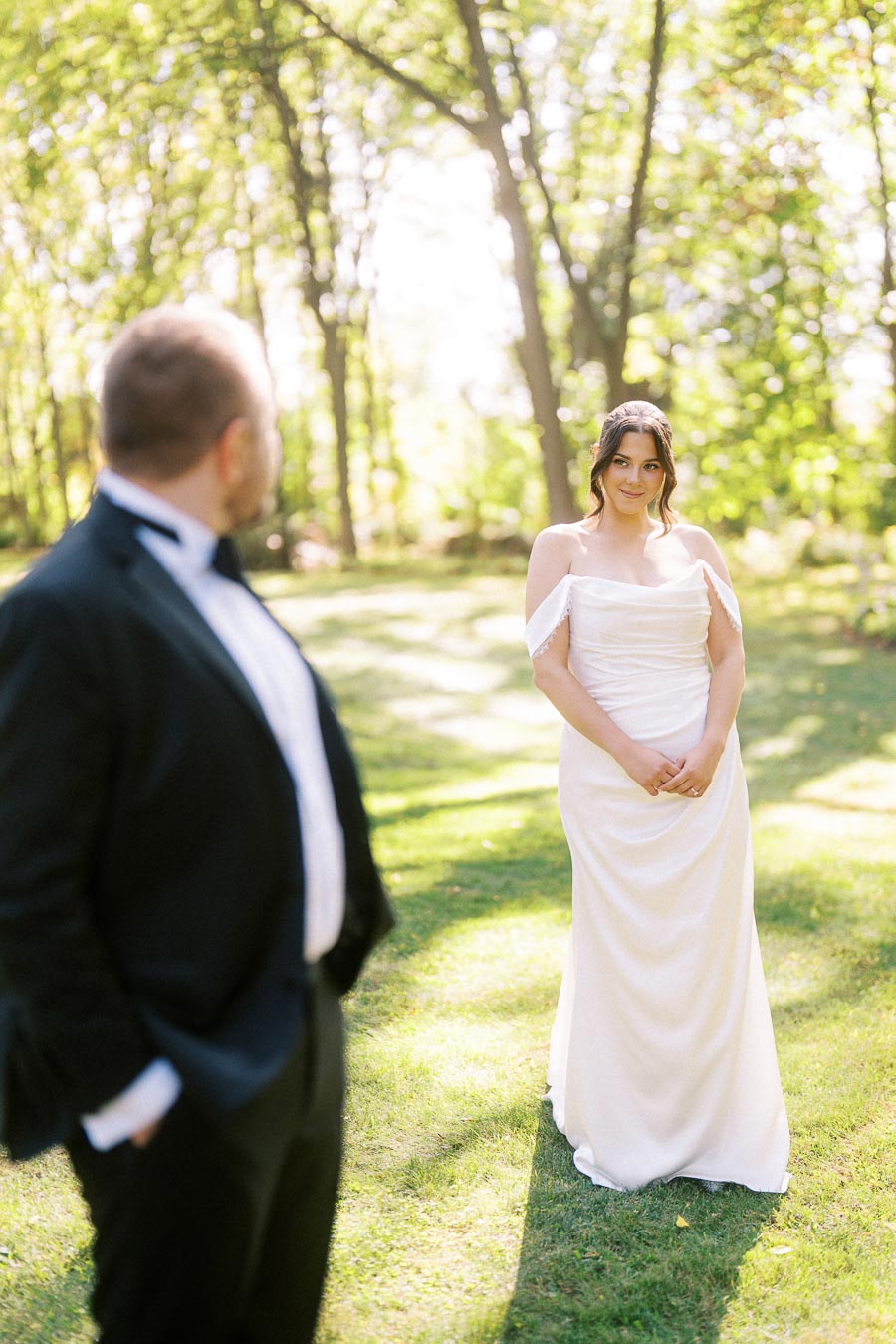 A bride in a white wedding dress stands in a sunlit, grassy area with trees in the background, looking at a man in a black suit in the foreground, creating a romantic outdoor wedding scene.