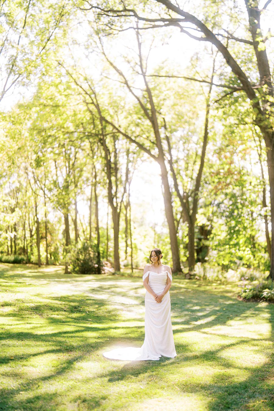 A bride in an elegant white dress stands serenely in a sunlit forest clearing, surrounded by tall trees and lush greenery, casting playful shadows on the grass.